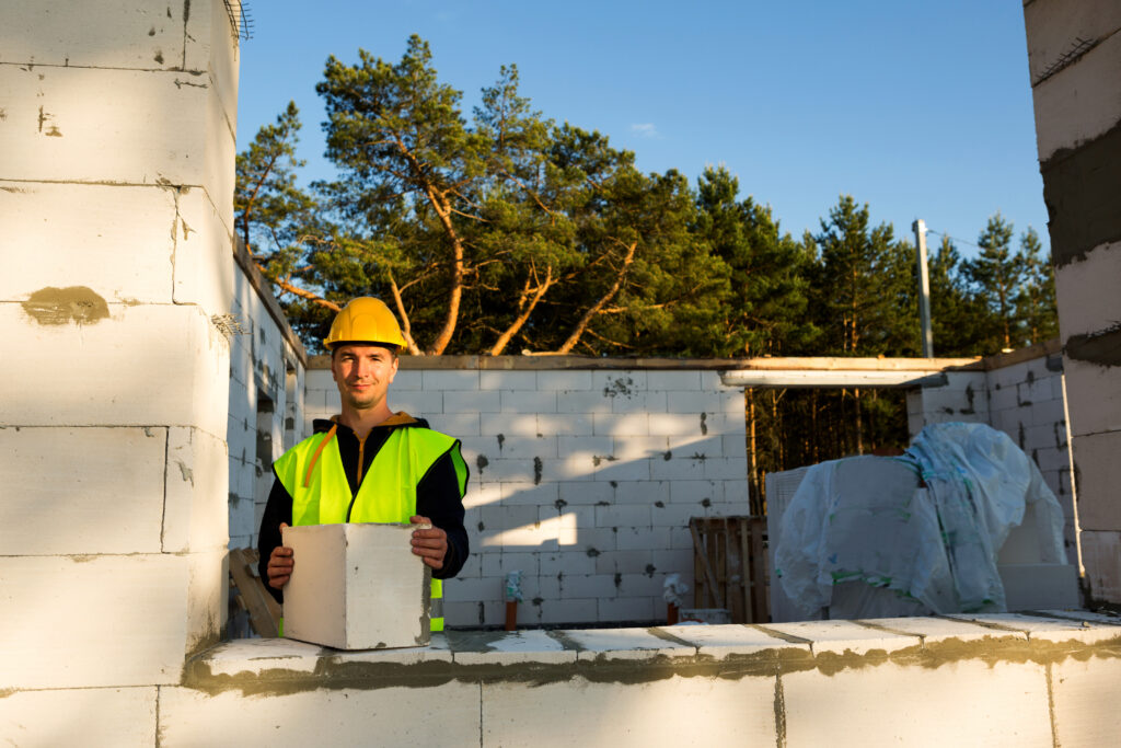 The builder holds a block of cellular concrete in his hands - the masonry of the walls of the house. Construction workers in protective clothing-a hardhat and a reflective vest.